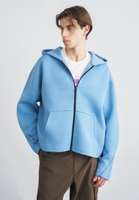 Φωτεινό γαλάζιο φούτερ με φερμουάρ από απαλό ύφασμα, με μεγάλο μπροστινό τσέπη και ρυθμιζόμενο κουκούλα. Συνδυάζεται με καφέ παντελόνι cargo.