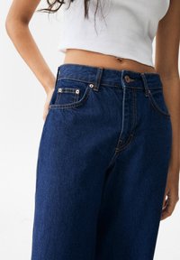 Jeans de mezclilla de talle alto en azul oscuro con costuras naranjas, que cuentan con bolsillos frontales y cierre de botón. Textura suave, corte de pierna ancha.