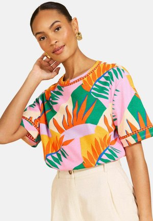 Kobieta z złotymi kolczykami w kolorowej koszuli w tropikalny wzór z krótkim rękawem i jasnobeżowych, wysokich spodniach, pozująca z dłonią przy twarzy.