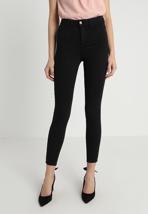 Mujer que lleva vaqueros negros pitillo de talle alto combinados con tacones negros de punta afilada y una camiseta sin mangas rosa claro.