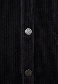 Tejido de pana negro con costillas verticales, con botones de metal plateado con detalle de logo y costuras de una sola puntada a lo largo de los bordes.