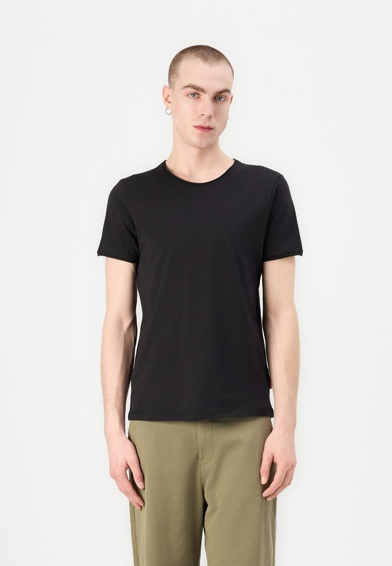Black cotton T-shirt with a crew neck, short sleeves, and a relaxed fit. Model wears olive green pants. Simple design with no patterns.
