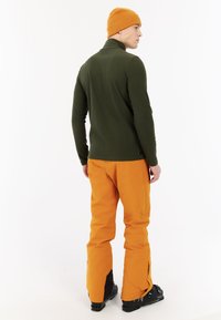 Maglia a maniche lunghe verde oliva abbinata a pantaloni arancioni. Il modello indossa un berretto di maglia arancione, mostrando stivali neri con dettagli blu.