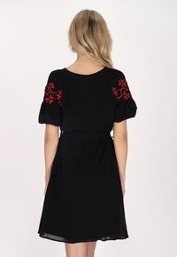 Schwarzes Kleid mit kurzen Puffärmeln, verziert mit roter floralem Stickerei auf den Schultern, tailliertem Bund und fließendem Rock. Weicher Stoff und lässiger Schnitt.