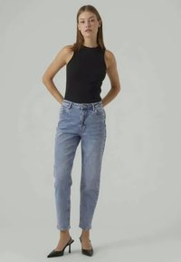 Črna brezrokavnica v kombinaciji z modrimi visokim pasom jeans. Model nosi črne尖toed čevlje; minimalen dizajn in gladke teksture.