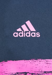 Fond textile bleu foncé avec un logo Adidas rose bien en vue, comportant trois bandes diagonales et une texture lisse.
