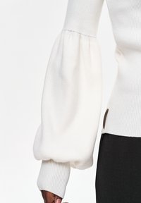 Witte geribbelde gebreide trui met volumineuze, geplooide ballonmouw en aangesloten manchetk, gedragen met zwarte broek aan een donkergekleurde arm.