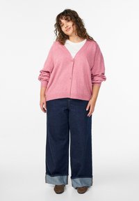 Rozā adīta kardigāna ar V-veida dekoltē un zelta pogu aizdari, kombinēta ar platām denima biksēm ar atlocītu apakšmalu un brūnām kurpēm.