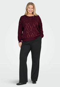 Burgundowy sweter z diamentowymi wycięciami, długimi rękawami i luźnym krojem, w połączeniu z czarnymi spodniami z wysokim stanem i czarnymi butami.