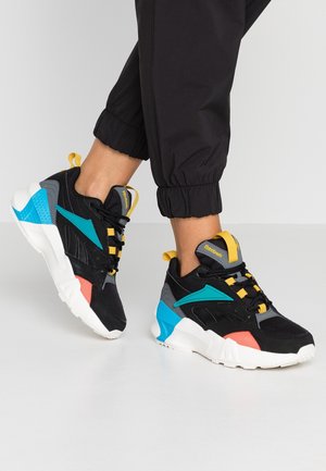Persona que lleva zapatillas Reebok negras, teal, coral, gris y amarillas con suelas blancas y pantalones deportivos negros sobre un suelo blanco.