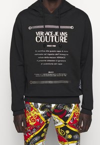Black hooded sweatshirt featuring gold text and rope graphics, paired with colorful patterned pants in red, white, and gold accents.