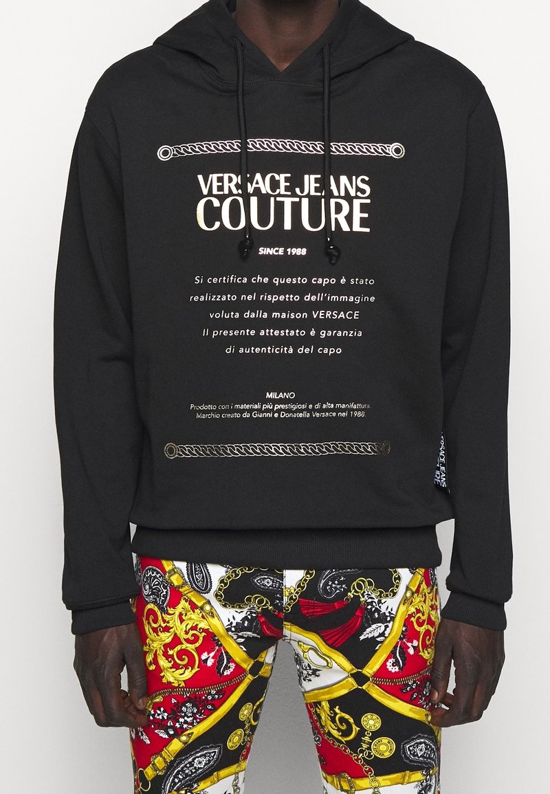 Black hooded sweatshirt featuring gold text and rope graphics, paired with colorful patterned pants in red, white, and gold accents.