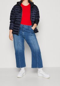 Marineblaue Steppjacke, rotes geripptes Oberteil, weit geschnittene blaue Jeans und weiße Turnschuhe. Das Outfit zeichnet sich durch eine Mischung aus Texturen und geschichteter Kleidung aus.