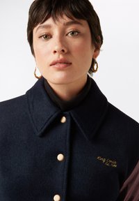 Marinblå kavaj i ullblandning med skjortkrage, träknappar och guldig broderad logotyp. Kavajen har kontrasterande mörka ärmar.