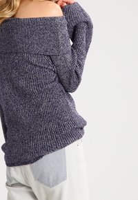 Femme portant un pull violet en maille à épaules dénudées et un jean en denim clair, vue de dos sur un fond uni.