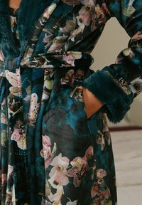 Floral patterned robe with teal faux fur collar and cuffs, tied waist belt, and hand tucked into side pocket.