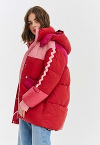 Žena v červené a růžové prošívané bundě s kapucí a vlnitým vzorem na rukávě, v kombinaci s modrými džíny, na jednobarevném bílém pozadí.