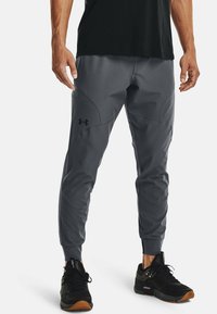 Muž v šedých sportovních kalhotách Under Armour a černých teniskách, stojící s uvolněnými rukama podél těla na bílém pozadí.