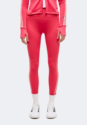 Άτομο φορά φωτεινά κόκκινα κολάν Adidas με ροζ ρίγες στο πλάι, λευκές κάλτσες, λευκά αθλητικά παπούτσια Adidas και ένα ταιριαστό κόκκινο μπουφάν.