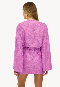 Robe mini texturée violette avec manches longues et taille froncée. Motif floral sur l'ensemble et finition à franges le long des bords.