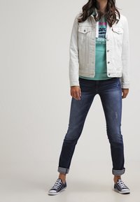 Giacca di denim bianco sopra un felpa color teal, jeans blu scuro arrotolati e sneakers navy, con dettagli scoloriti sui jeans.