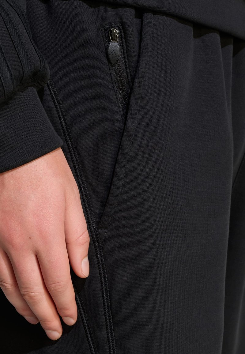 Schwarze Sporthose mit Reißverschlusstasche und strukturierten Bündchen, mit Nahaufnahme, die glattes Gewebe und Nähte hervorhebt.