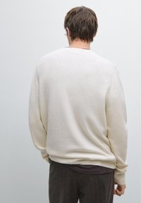 Krämfärgad tröja med rund halsringning, ribbade manschetter och fåll, samt en mjuk textur; bärs över en mörk skjorta, sedd bakifrån.