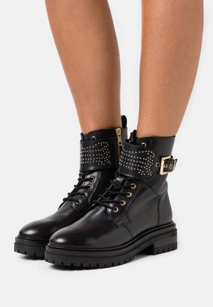 Mustat nahkaiset nilkkurit, joissa on matala korko, kullanväriset yksityiskohdat, niititty varsi ja nauhoitus. Sivuvetoketju helppoa pukeutumista varten.