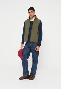 Grøn vest over en mørkeblå trøje, blå jeans og brune sko. Holder en rød kasket. Glat stof, afslappet pasform og lynlås foran.