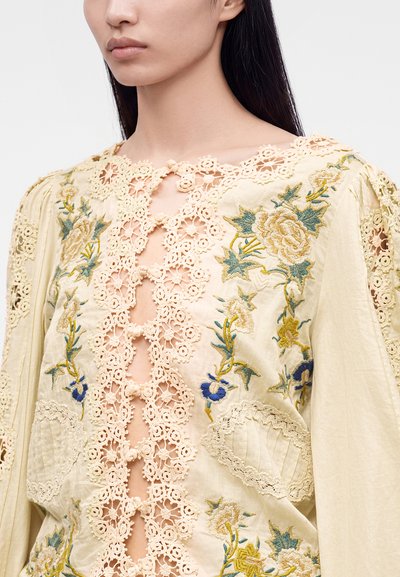Woman wearing cream blouse with floral embroidery and lace detailing along the front opening and shoulders.
