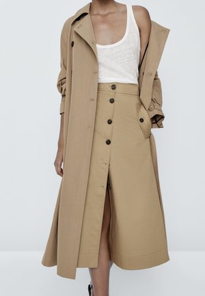 Woman wearing beige trench coat over buttoned beige skirt and white sleeveless top, standing against plain background.