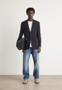 Blazer azul-marinho com bainhas brancas, usado sobre uma t-shirt branca, combinado com jeans de perna reta azul-claro e ténis cinzentos, segurando uma mala preta.