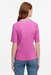 Kurzarm-Poloshirt in leuchtendem Pink mit gemustertem Kragen. Der Stoff wirkt glatt; das Shirt hat eine lockere Passform und reicht bis zur Hüfte.