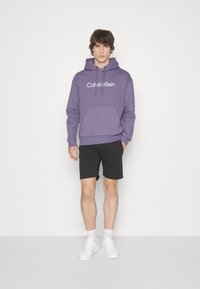 Felpa con cappuccio viola e tasca frontale a marsupio, con logo "Calvin Klein" bianco. Indossata con pantaloni corti neri e sneakers bianche.