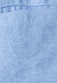 Lys blå denimstof med en let struktureret overflade, der har synlige sømme langs den øverste kant og en glat finish.