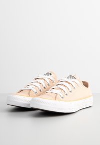Beiga canvas sneakers med vita snören, gummisulor och gula accenter nära sulan. Har en logotyp patch på tungan och prickig sula.