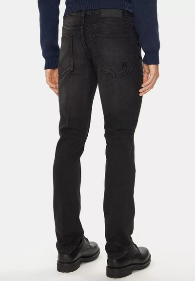 Jeans in denim nero con una vestibilità slim, caratterizzati da tasche posteriori, cuciture sottili e una superficie testurizzata. Indossati con stivali neri stringati.