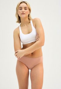 Weißes geripptes Croptop kombiniert mit hellrosa nahtlosen Boxershorts. Das Model hat die Arme verschränkt und zeigt die Texturen und Passform des Stoffs.