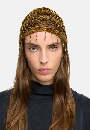 Kobieta nosząca złotą sieciowaną ozdobę na głowę z wiszącymi koralikami, długie brązowe włosy i czarny top z wysokim kołnierzem na jednolitym tle.