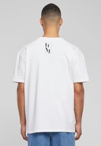 Hvid bomulds t-shirt med løs pasform, der har et lille sort grafisk design på den øverste ryg og korte ærmer. Bæres med blå denim shorts.
