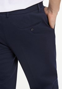 Pantalones navy fabricados en una tela suave, con un bolsillo lateral con cierre de botón y un corte ajustado. Sin patrones ni detalles de textura visibles.