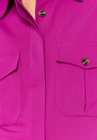 Bright pink shirt with two front pockets, vertical stitching, and a pointed collar. Features a button closure with a glossy black button.