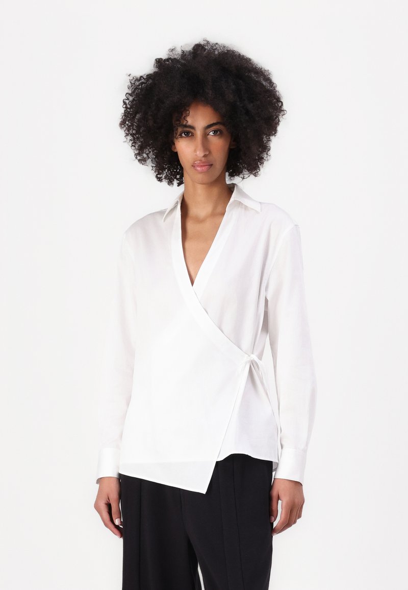 Blusa avvolgente bianca in tessuto leggero, con scollatura a V profonda, maniche lunghe e dettaglio con fiocco sul lato, abbinata a pantaloni neri.