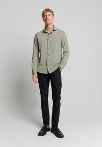 Olijfgroene, lange mouwen shirt met een knopenlijst aan de voorkant, zwarte knopen en een relaxed fit. Gepaarde met zwarte broek en zwarte schoenen.