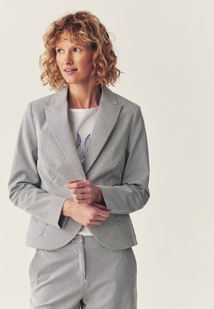 Kvinde med krøllet hår iført en hvid og navy pinstripe dragt og hvid skjorte med blåt blomsterdesign, der kigger til venstre.