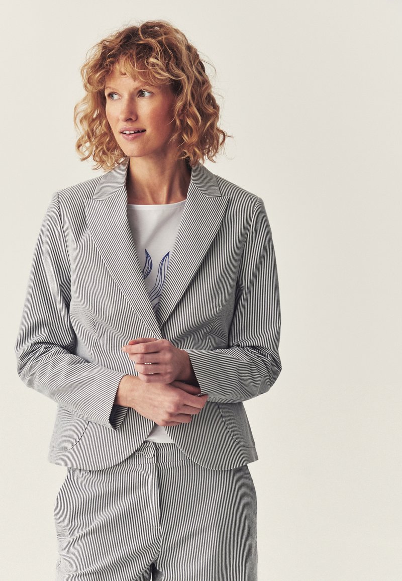 Femme aux cheveux bouclés portant un costume blanc à fines rayures bleu marine et une chemise blanche avec un motif floral bleu, regardant vers sa gauche.