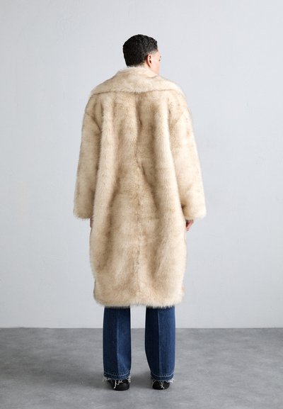 Šviesiai ruda dirbtinė kailio striukė su plačiais atlapais, ilgomis rankovėmis ir iki kelių siekiančiu dizainu. Derinama su mėlynomis platėjančiomis džinsinėmis kelnėmis ir juodais batais. Sklandi tekstūra.