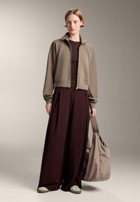 Tajtat jeansjacka i taupe, med dragkedja fram; matchas med flödande bordeauxbyxor. Håller en mjuk, ljusbrun väska; beige sneakers.