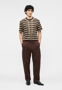 Jeune homme portant une chemise marron rayée, un pantalon marron texturé et des chaussures en cuir noir, debout les mains dans les poches sur un fond blanc.