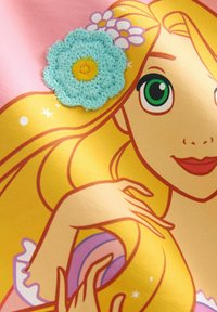 Ilustrácia tváre postavy s blond vlasmi, veľkými zelenými očami, oblečená v ružovo-žltej súprave, zdobená modrým háčkovaným kvetom.